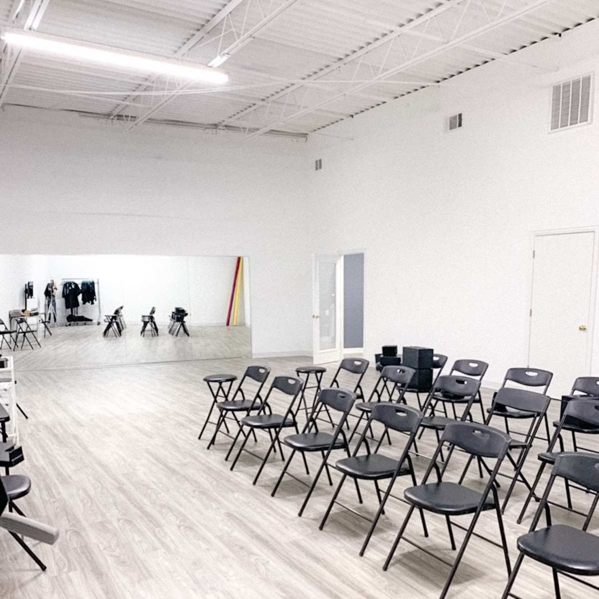 A spacious room with rows of black folding chairs facing a large mirror on a white wall.