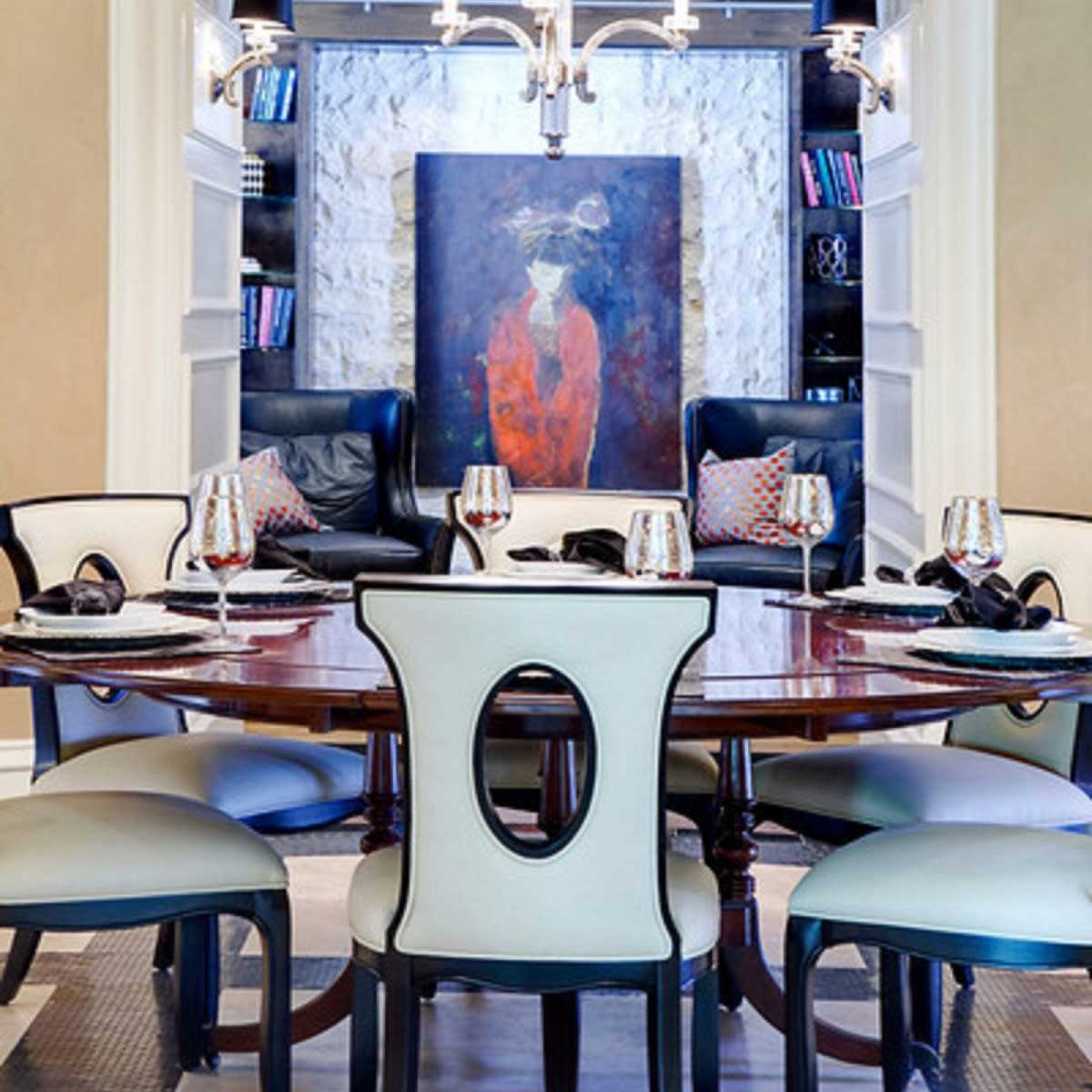 A dining room features a round table surrounded by upholstered chairs, a chandelier overhead, and a large painting displayed on the wall behind.