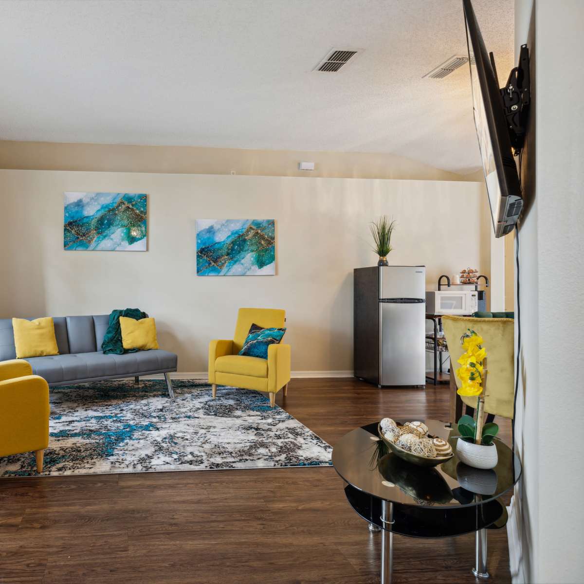 A living room features a gray sofa, two yellow chairs, decorative paintings on the walls, and a small refrigerator in the corner.