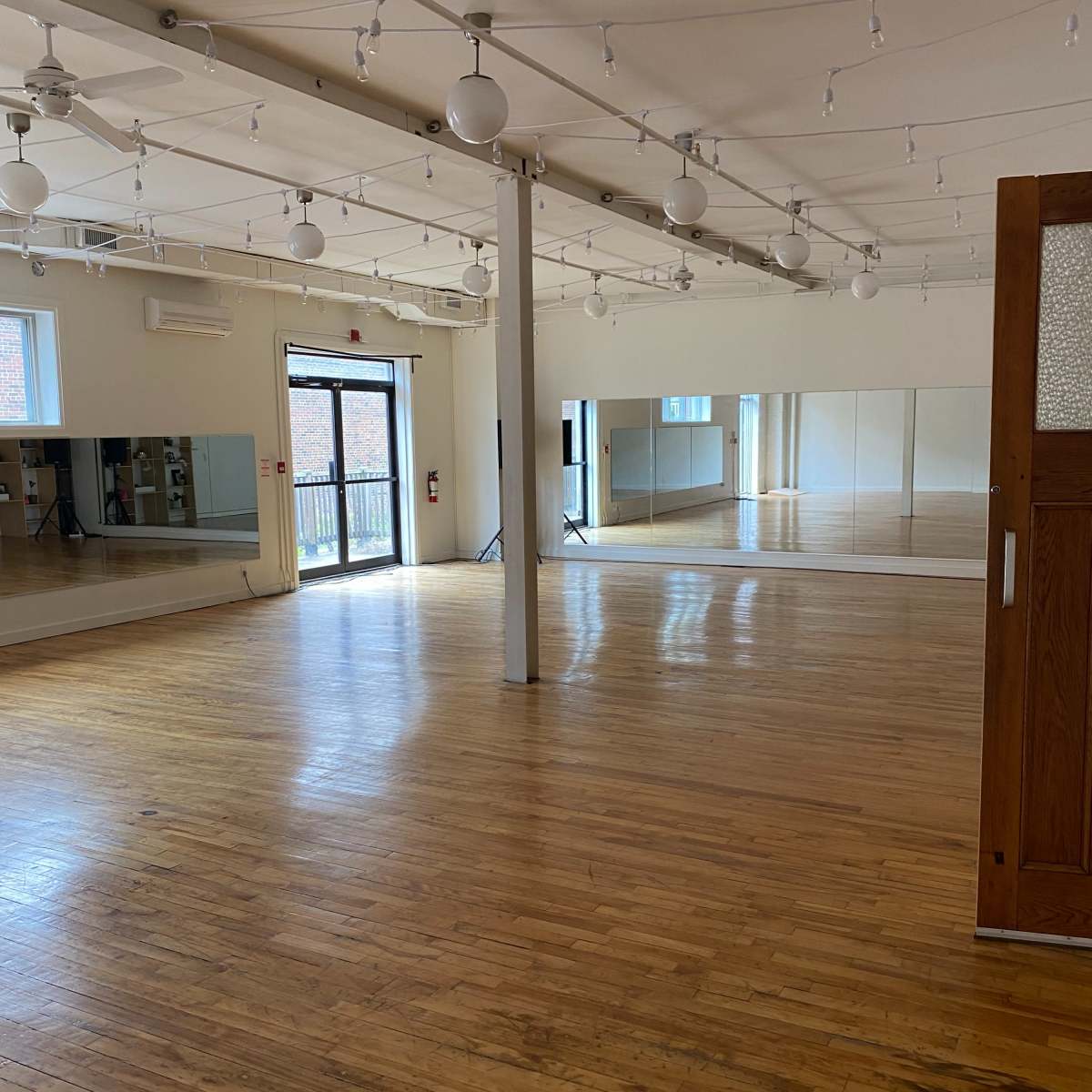 An empty dance studio with wooden floors, large mirrors along one wall, and natural light coming through a door leading outside.