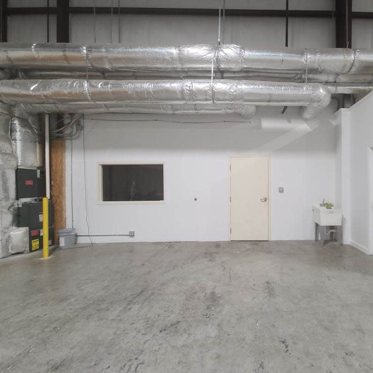 An empty industrial room with exposed metal ductwork, a door, and a small sink against a white wall.