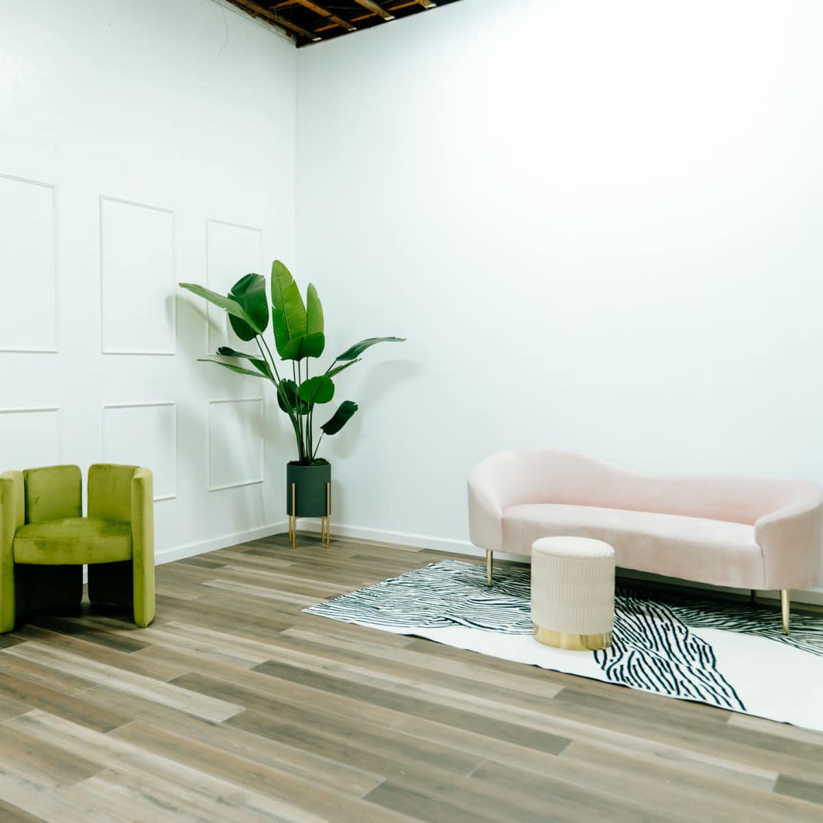 A minimalist interior with a pink couch, green armchairs, a small table, and a potted plant against a white wall with paneling.