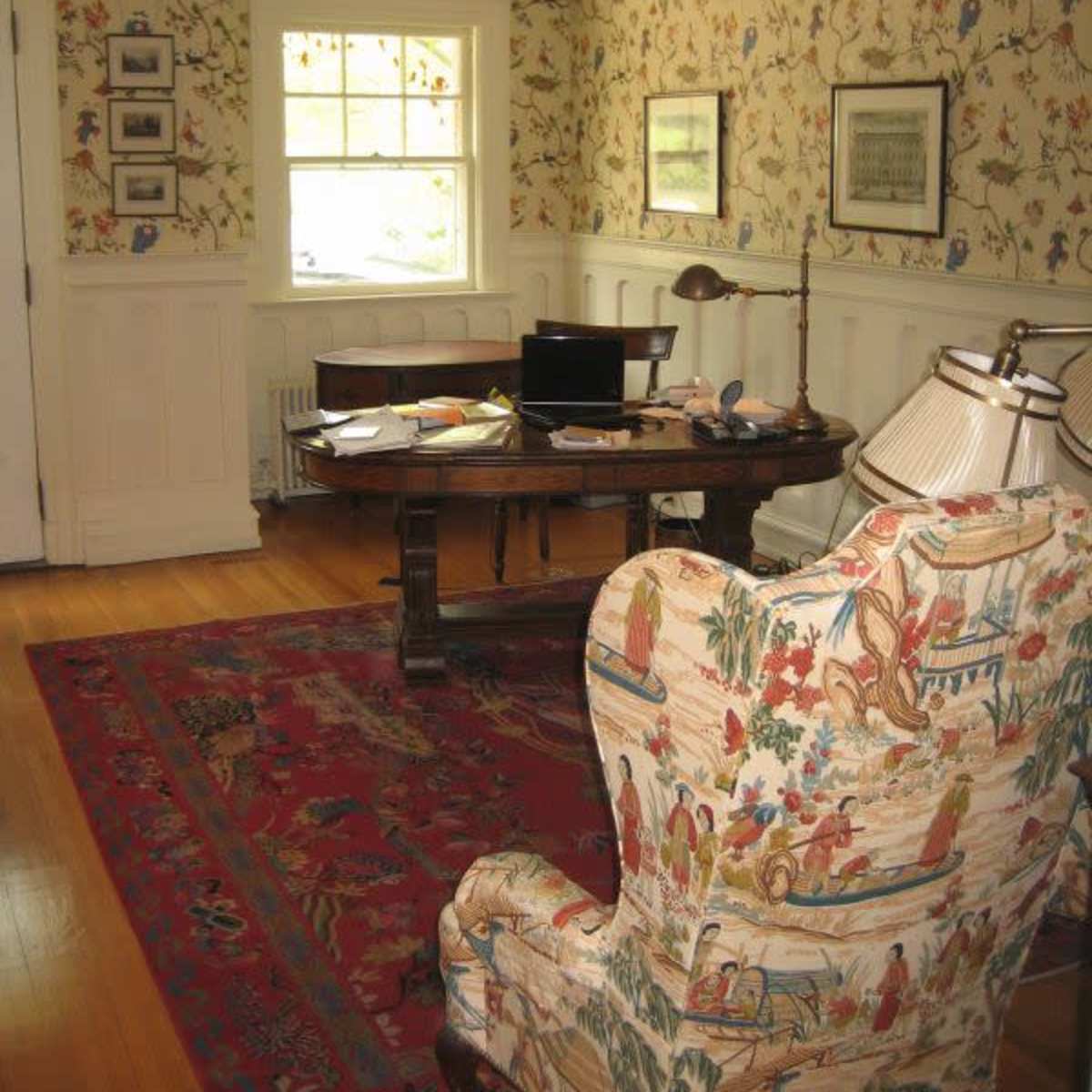 A well-lit home office with a wooden desk, a computer, and a large upholstered chair beside a patterned area rug.