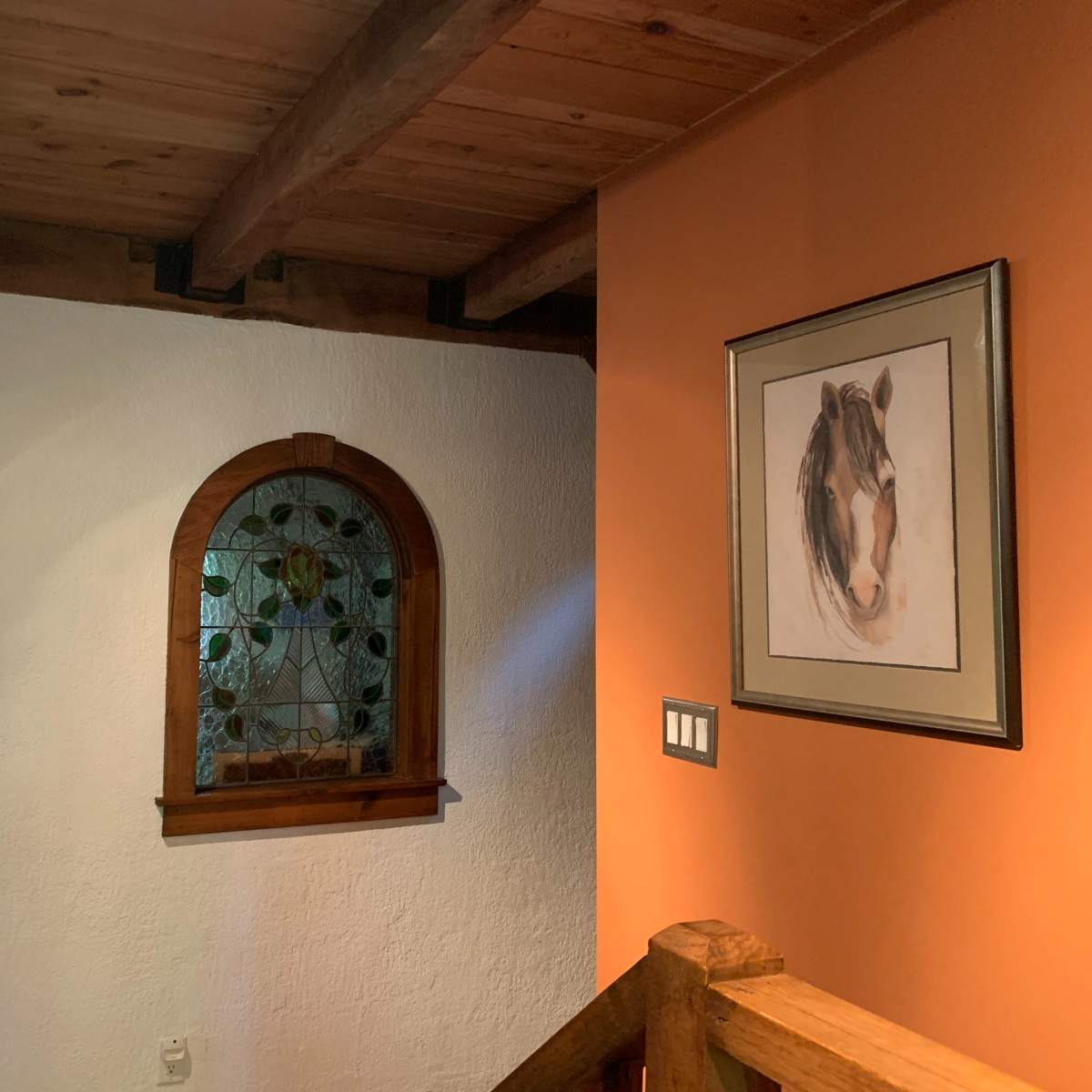 A corner of a room featuring an arched window with stained glass, a wooden railing, and a framed picture of a horse on an orange wall.