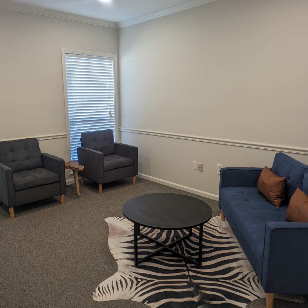 A living room with two gray armchairs, a blue sofa with decorative cushions, a round black coffee table, and a zebra-print rug.