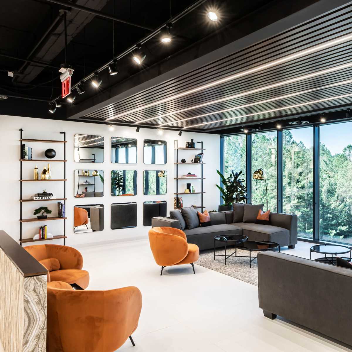 A modern lounge area with large windows, featuring gray sofas and orange armchairs arranged around a coffee table, alongside decorative shelves and greenery outside.