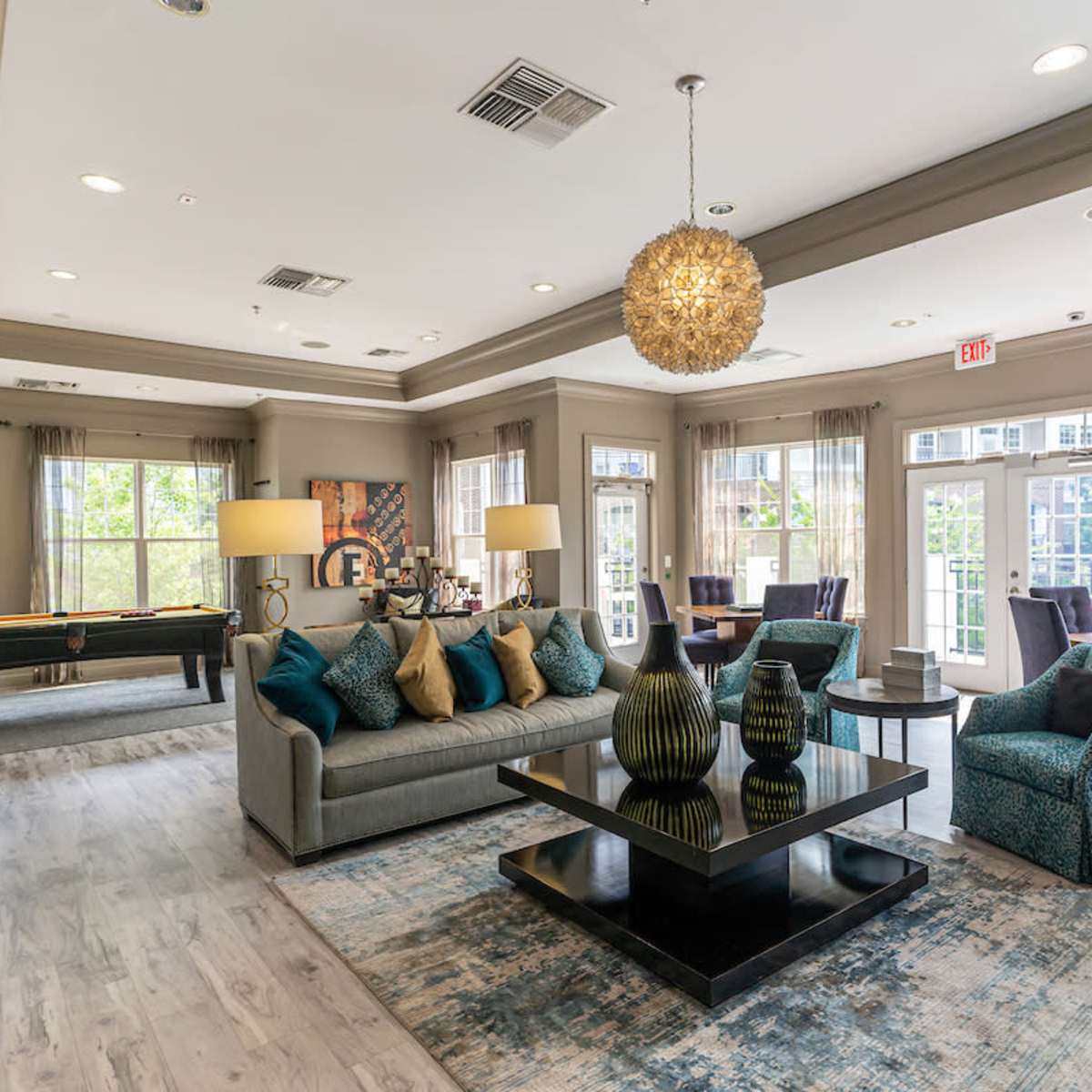 A spacious, modern lounge area featuring a large sofa, chairs, a coffee table, and a pool table, with abundant natural light from large windows.