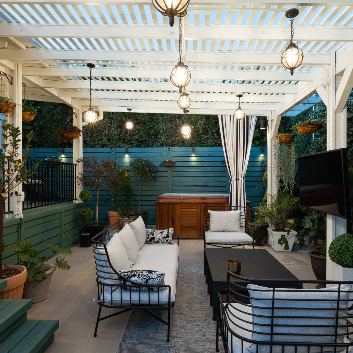 A covered patio features a seating area with a sectional sofa, a coffee table, and a television, surrounded by decorative plants and lighting.