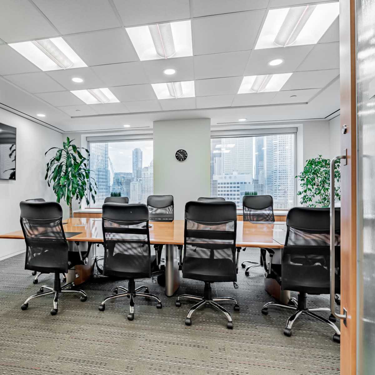 A conference room with a long wooden table and eight black chairs is visible through glass doors, featuring large windows that overlook a city skyline.