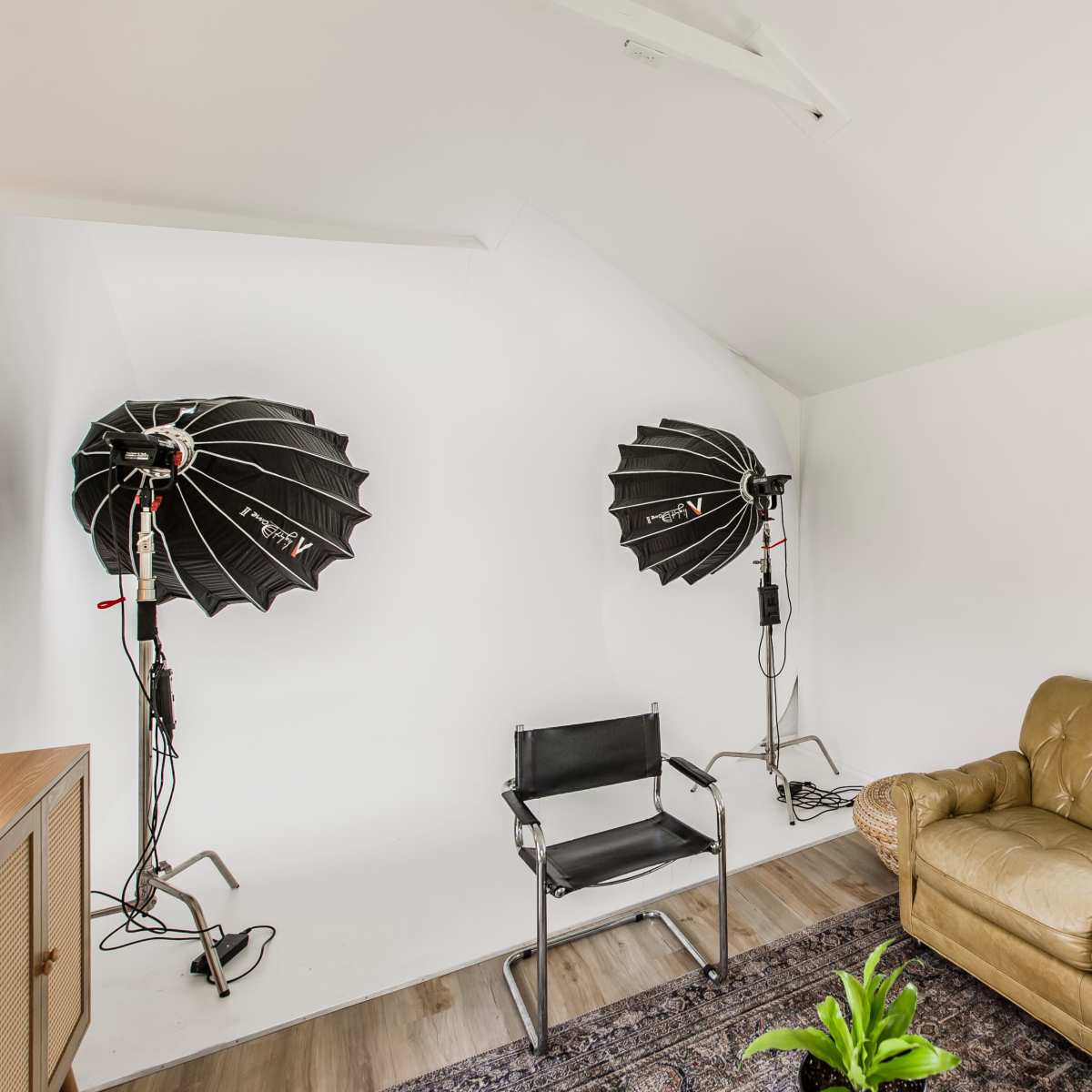 A well-lit room features two photography light setups and a tan leather sofa near a window.