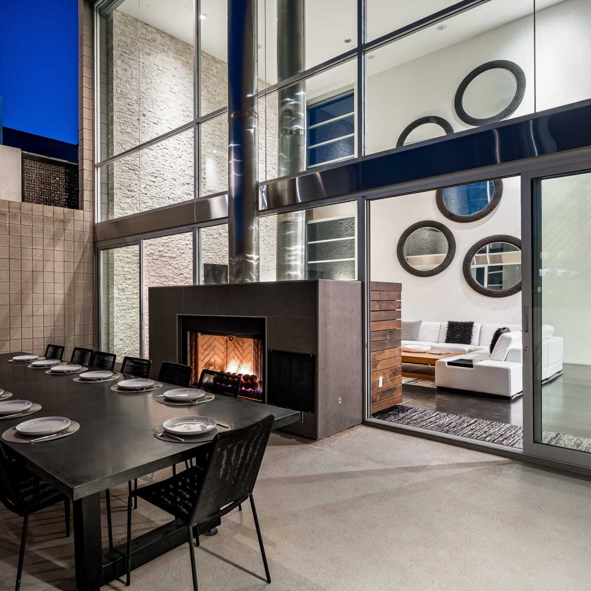 A modern outdoor dining area features a long table set for dinner next to a glass-enclosed living space with a fireplace and circular mirrors.