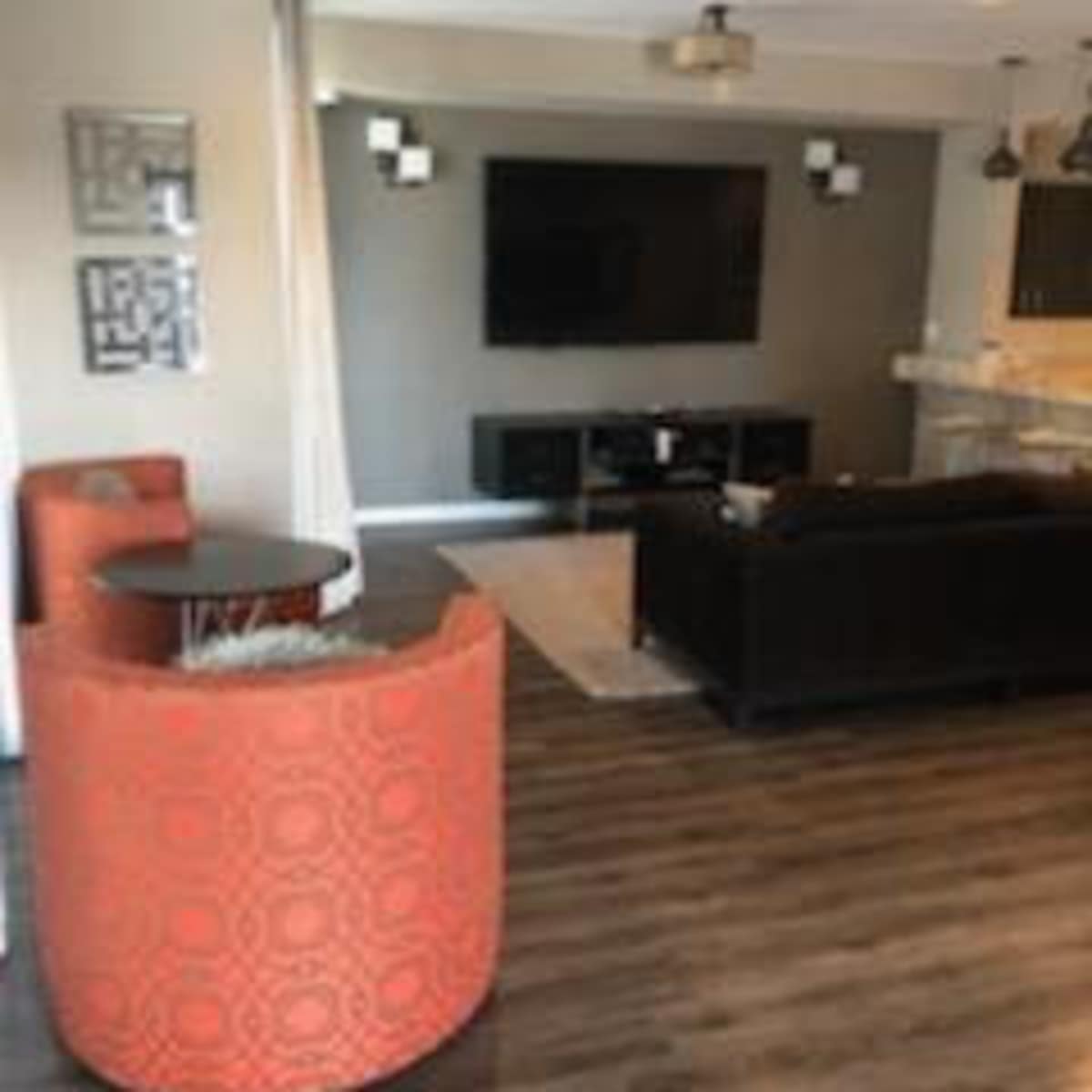 A modern living room with a television mounted on the wall, a dark sofa, an orange armchair, and a dining area with a bar counter.