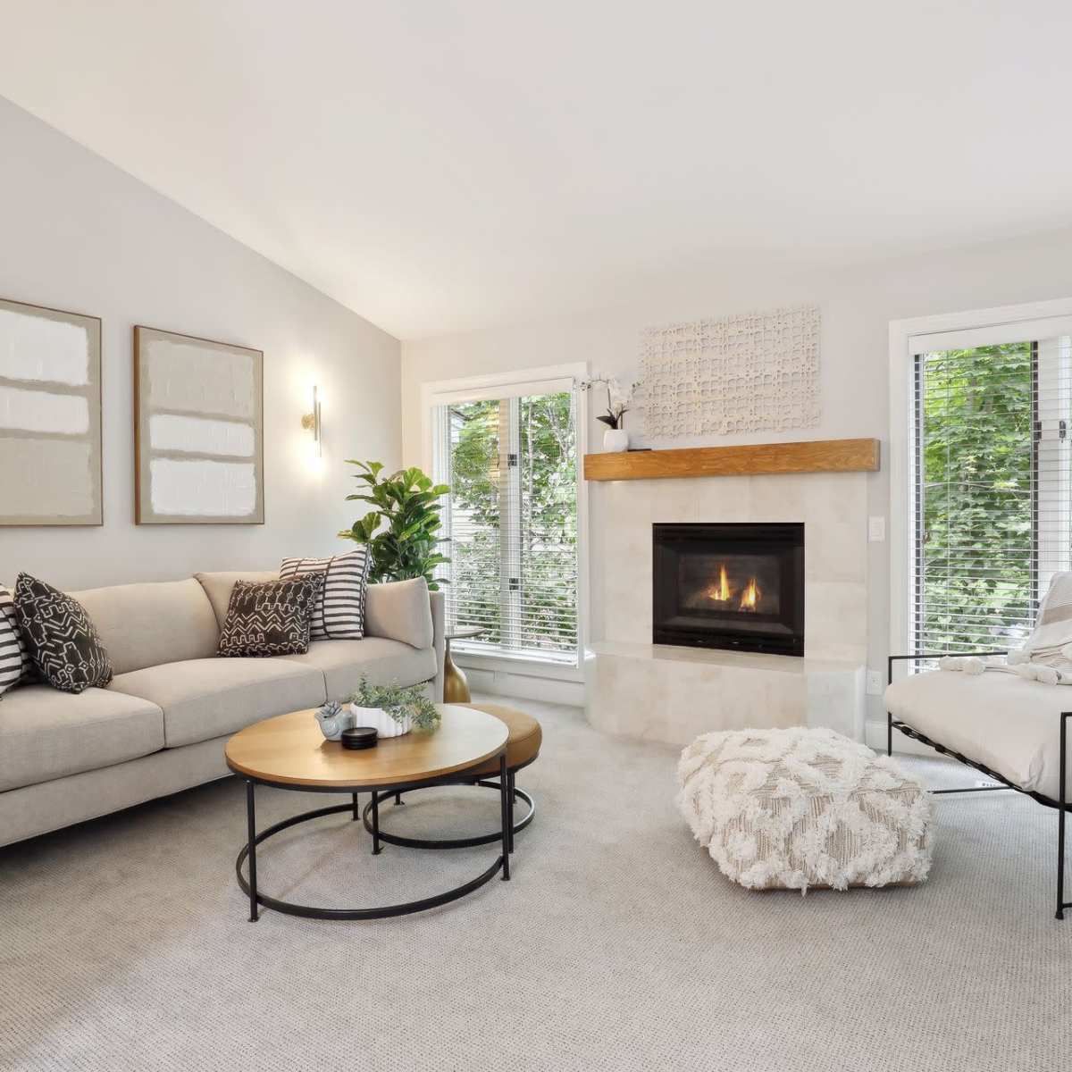 The living room features a beige sofa and an armchair facing a stone fireplace, with large windows revealing a view of greenery outside.