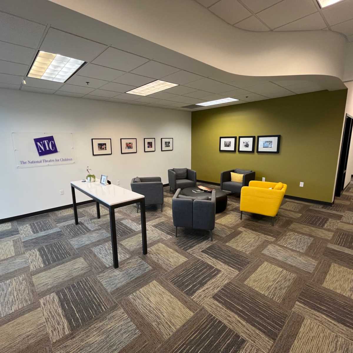 A modern office lobby with gray and yellow chairs arranged around a small table, framed pictures on the wall, and a logo banner on the left.