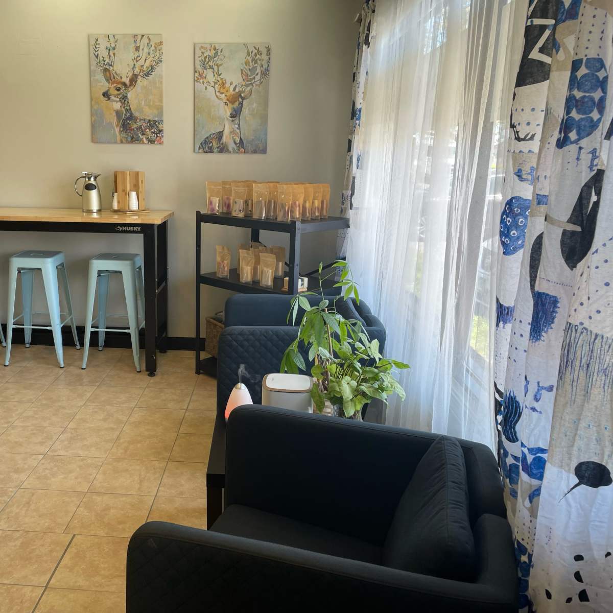 A bright, modern seating area with a long wooden table, blue stools, and two black couches, featuring decorative artwork and plants.
