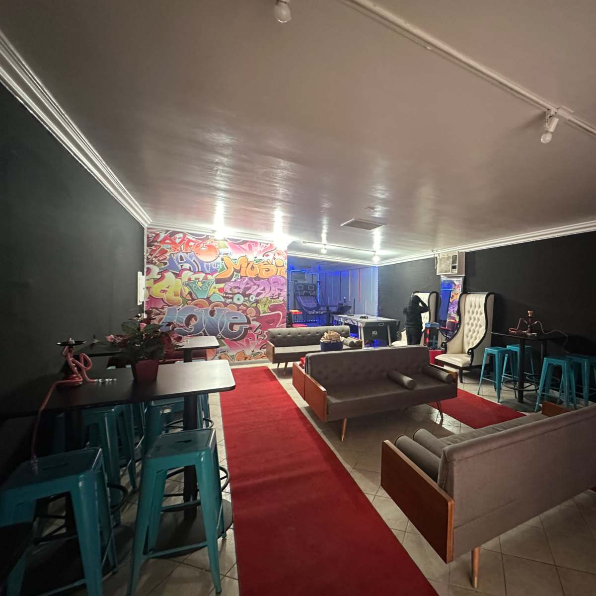 The interior features a lounge area with gray sofas, blue stools, and a graffiti-style accent wall under soft lighting.