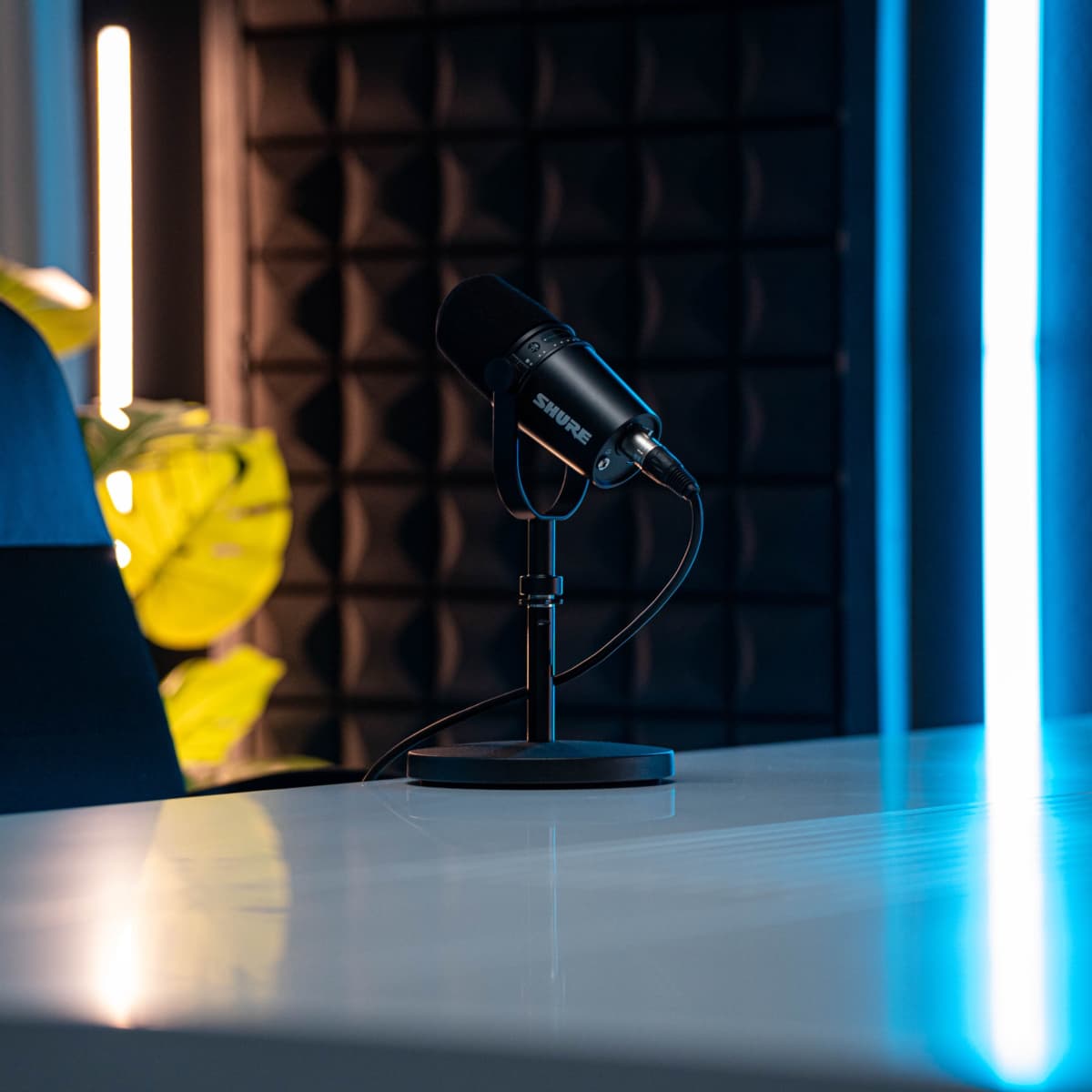 A black microphone stands on a sleek desk, with a textured black wall and dimly lit blue and white light fixtures in the background.