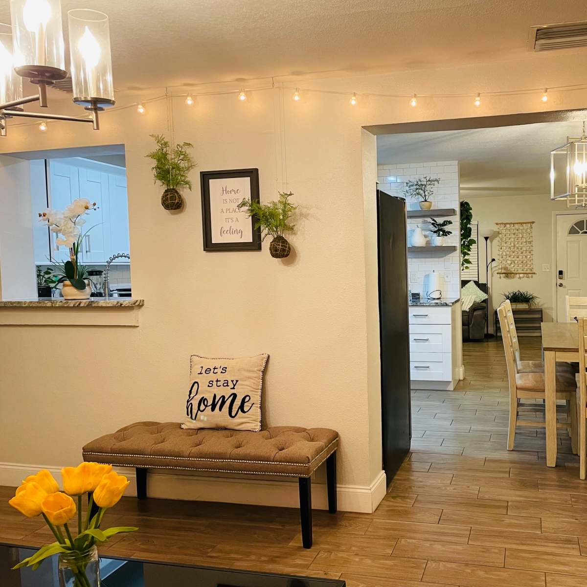 A cozy living area features a plush bench with a "let's stay home" pillow, leading to a dining space with a wooden table and chairs, adorned with warm string lights.