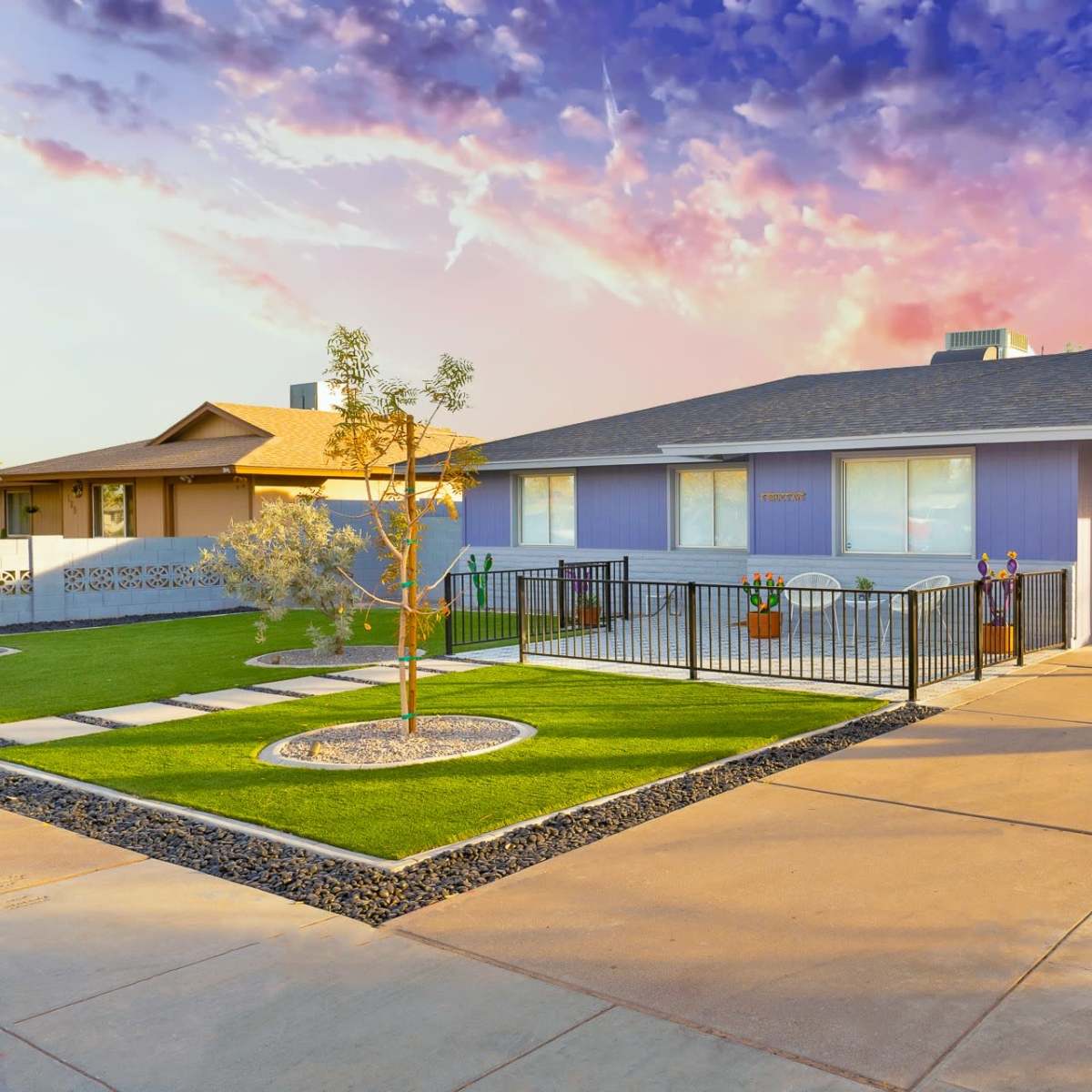 A single-story, lavender-colored house with a landscaped yard, bordered by a concrete driveway and a decorative fence, sits under a colorful sky.