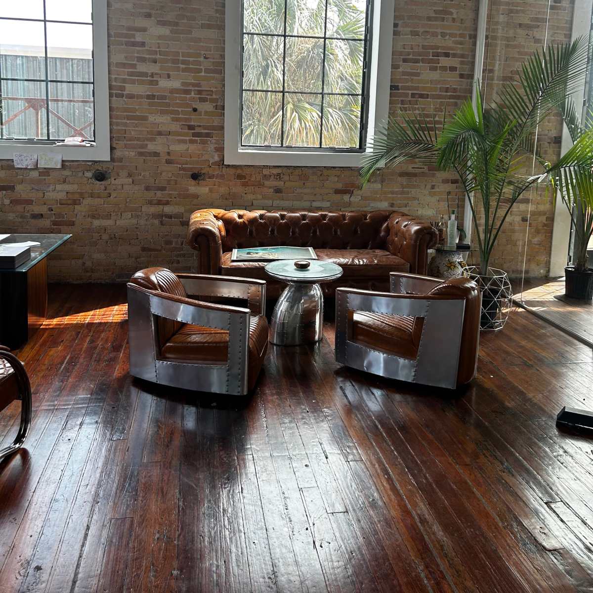The scene features a leather chesterfield sofa and two contemporary chairs surrounded by large windows and a palm plant in a room with exposed brick walls and polished wooden floors.