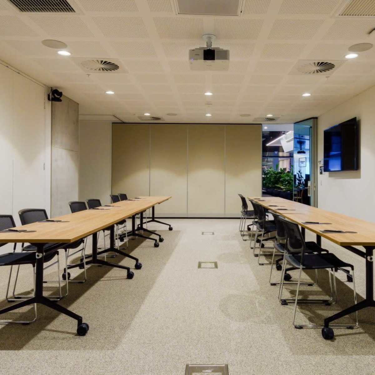 A modern conference room features a long wooden table with black chairs arranged on either side and two screens mounted on the walls.