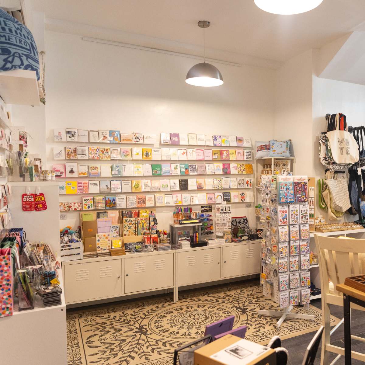 A cozy shop interior filled with greeting cards, stationery items, and assorted bags, with a patterned floor and shelving displaying various products.