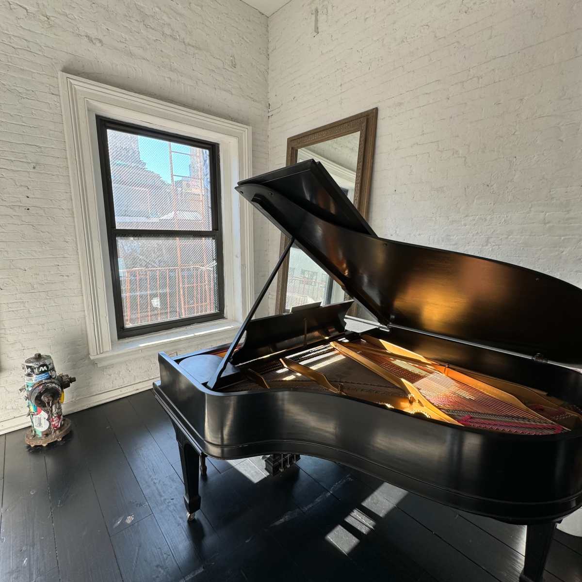 A grand piano is positioned in a sunlit corner of a room with exposed brick walls and a large window.