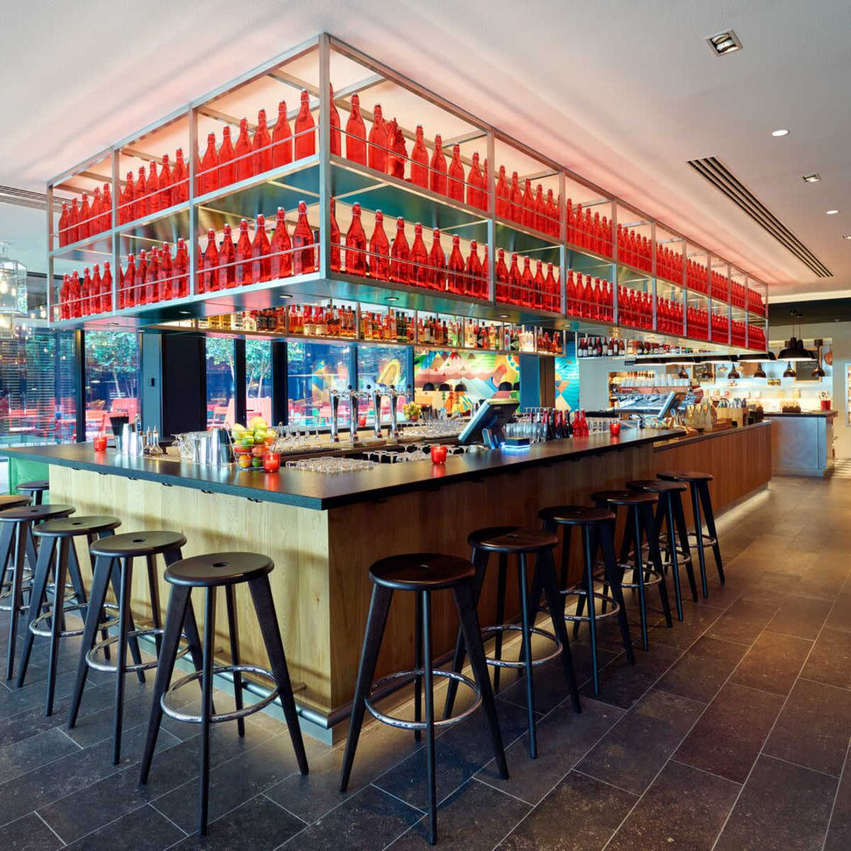 A modern bar with a long counter, black stools, and shelves filled with red bottles overhead, set in a stylish, open space.