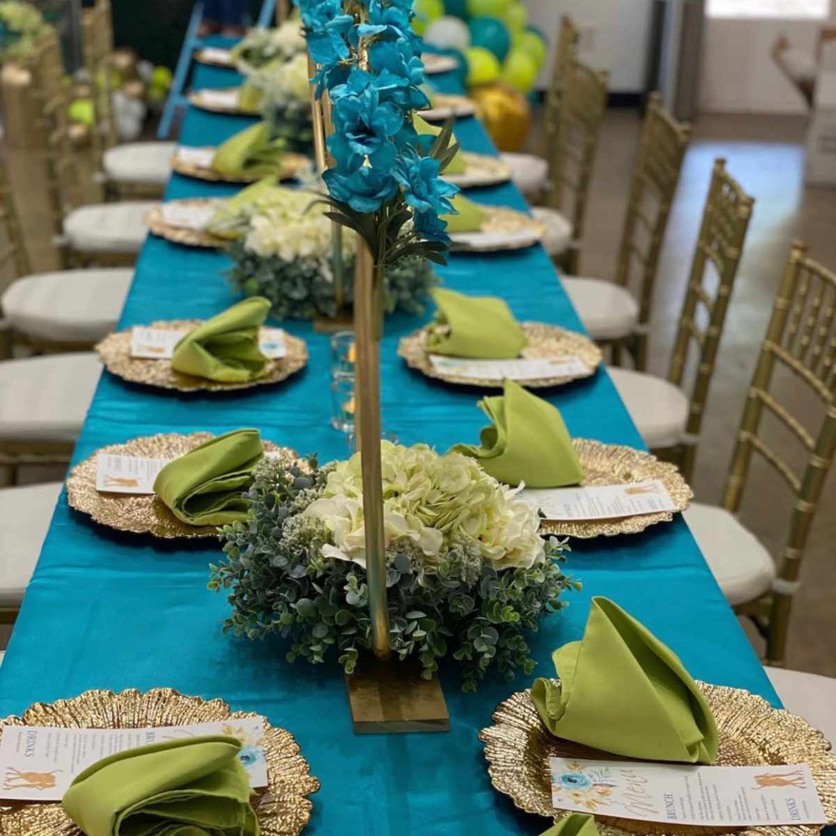 A long table set for a formal event features gold chargers, green napkins, and a floral centerpiece with blue orchids and white blooms.