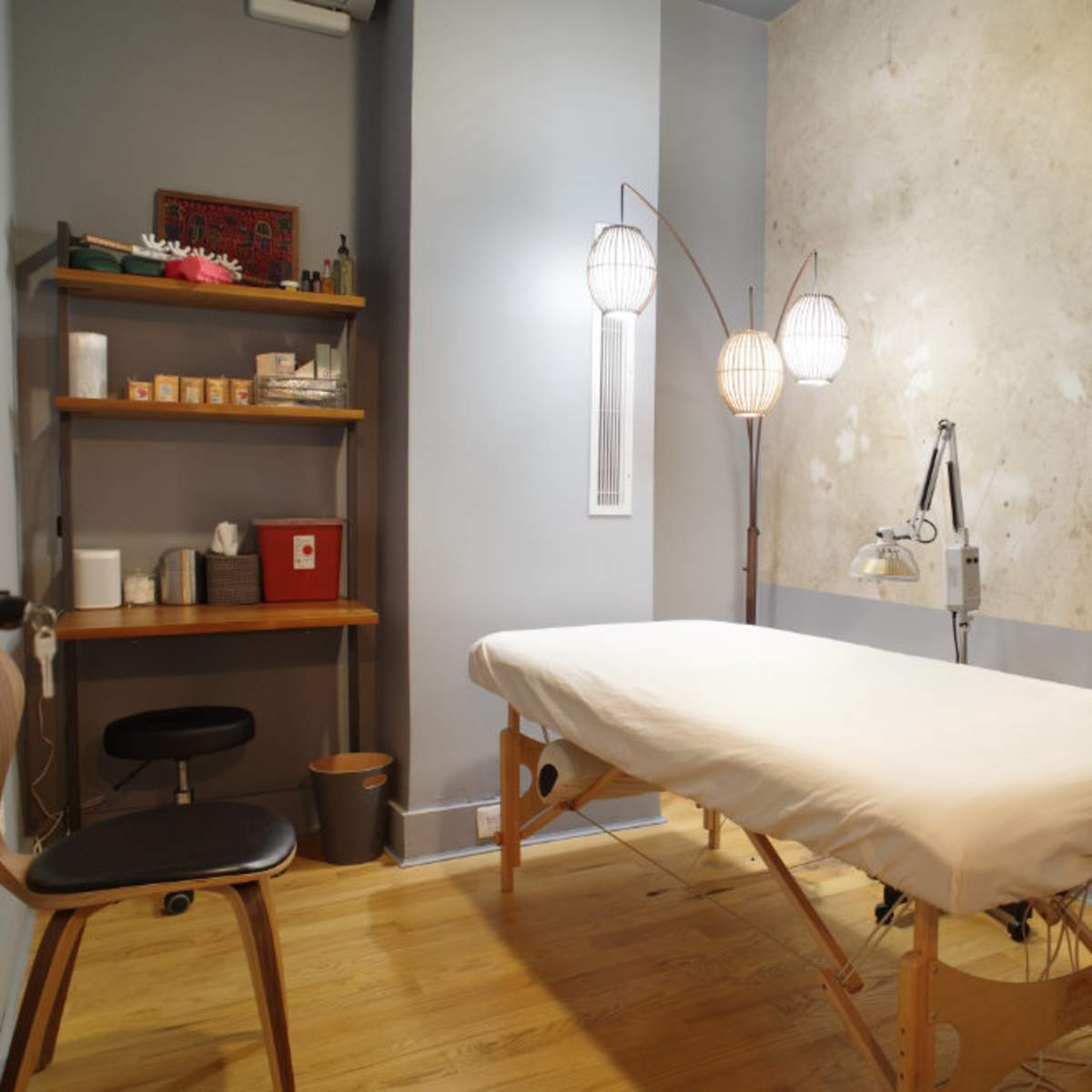 A massage room with a treatment table, a wooden shelf containing various items, and three floor lamps against a textured wall.