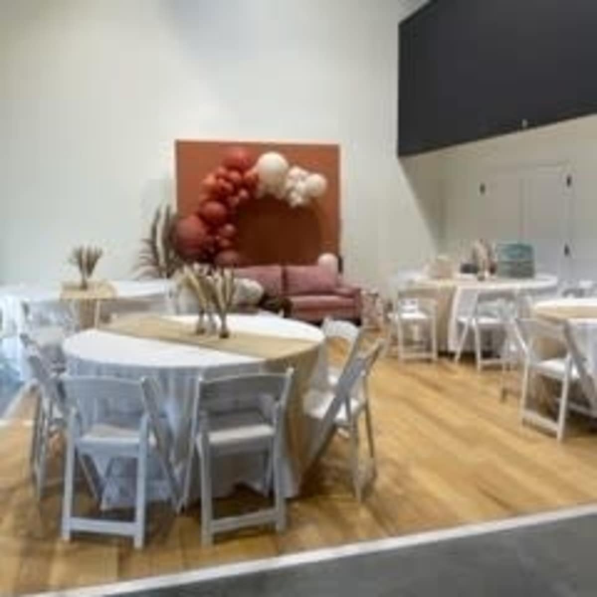 A spacious event venue with several round tables set up, white chairs, and a decorative wall featuring balloons and a couch in the background.