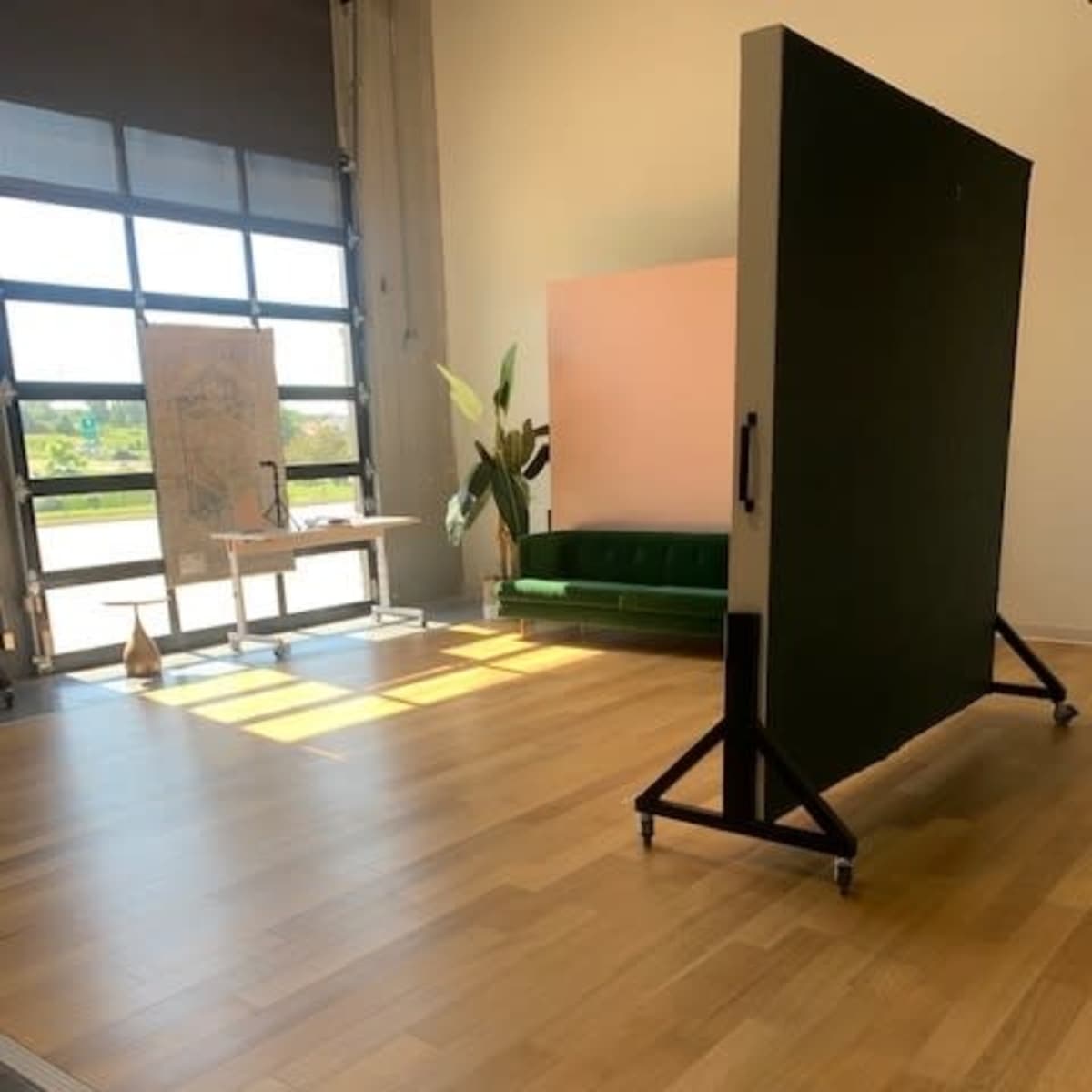 An indoor space featuring a green couch, a large movable panel on wheels, and a table with a standing drawing board, all illuminated by natural light from a large window.