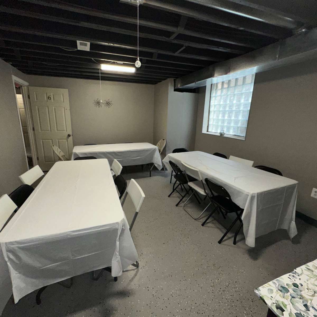 A basement room with several long tables covered in white tablecloths and chairs arranged around them, along with a mirror and a window featuring a glass block design.