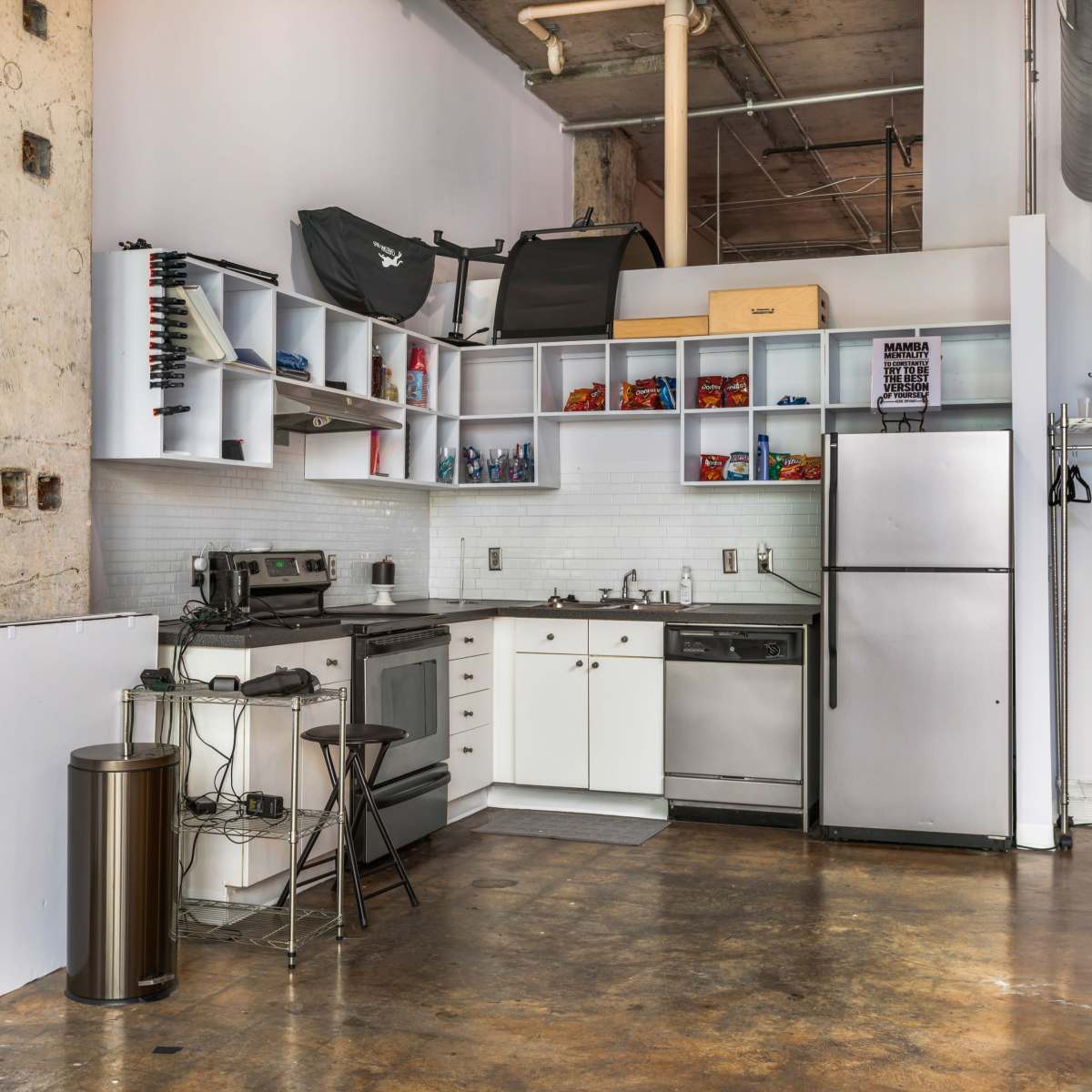 A compact kitchen area featuring a stove, oven, refrigerator, and shelving, with industrial-style walls and flooring.