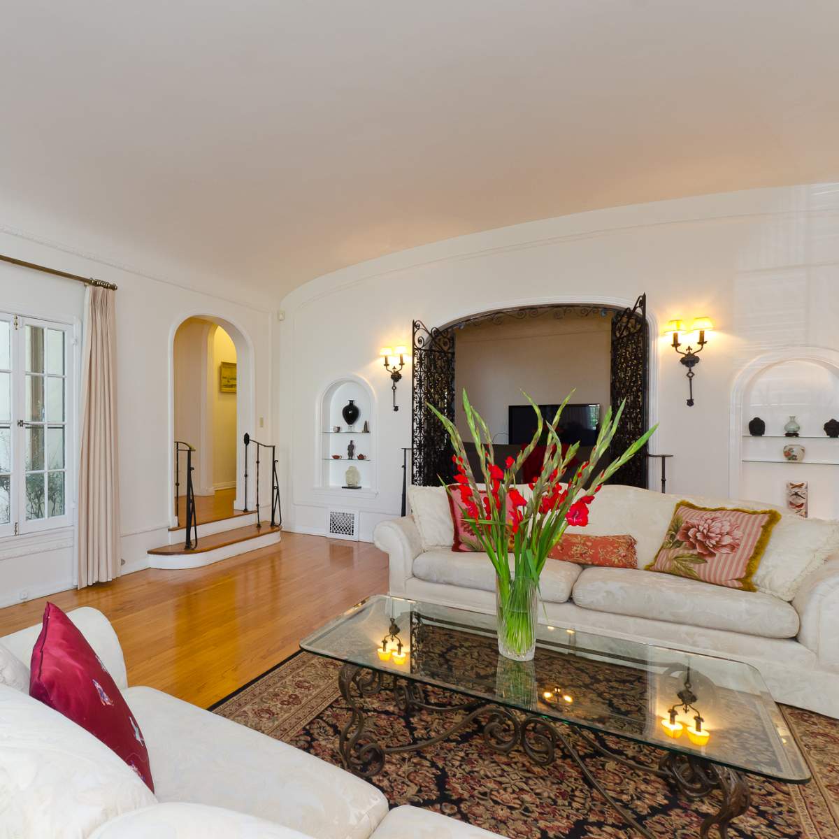 The scene features a bright living room with a white sofa, glass coffee table, and decorative plants, leading to an arched entryway with additional seating and wall-mounted shelves.