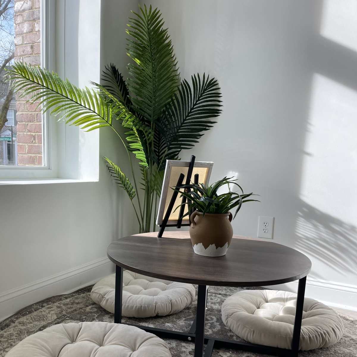A low wooden table is surrounded by three round, tufted floor cushions, with a potted plant and a photo frame on the table near a large window.