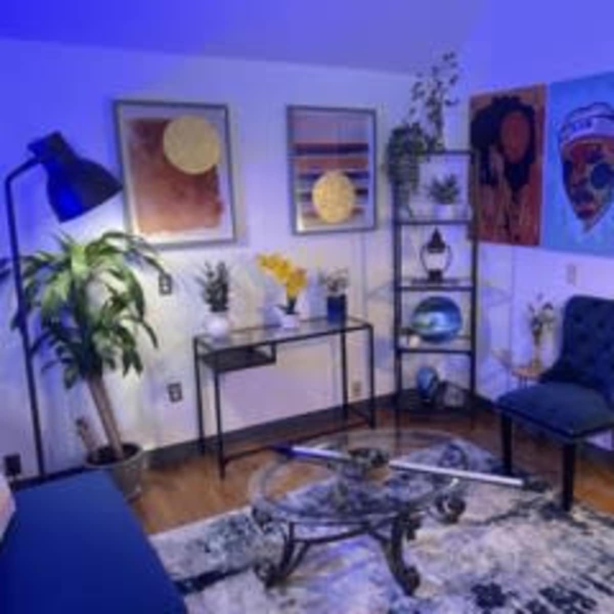 A modern lounge area featuring a blue armchair, a glass coffee table, and several pieces of framed artwork on the walls.