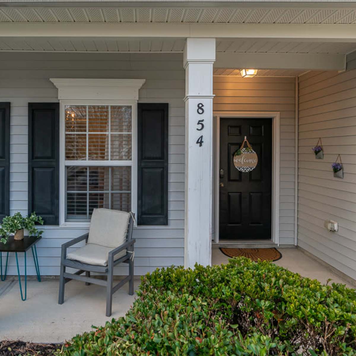 A front porch with two gray chairs, a small table, and welcoming decor on the door, framed by two white pillars.
