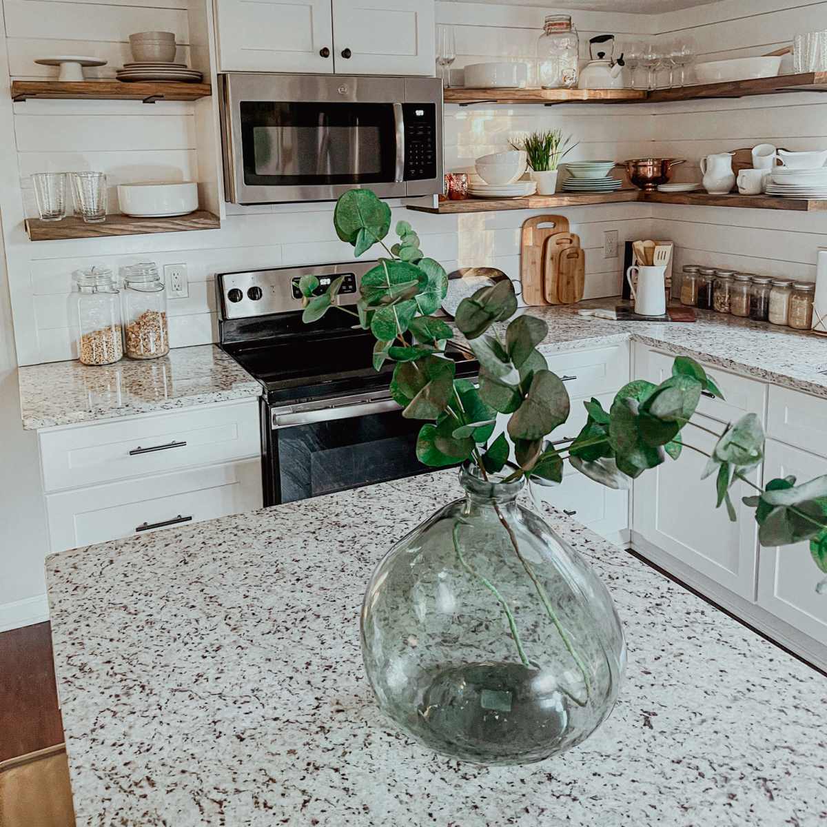 A modern kitchen features white cabinets, open shelving with various dishes and glassware, and a stone countertop with a decorative glass vase and greenery.