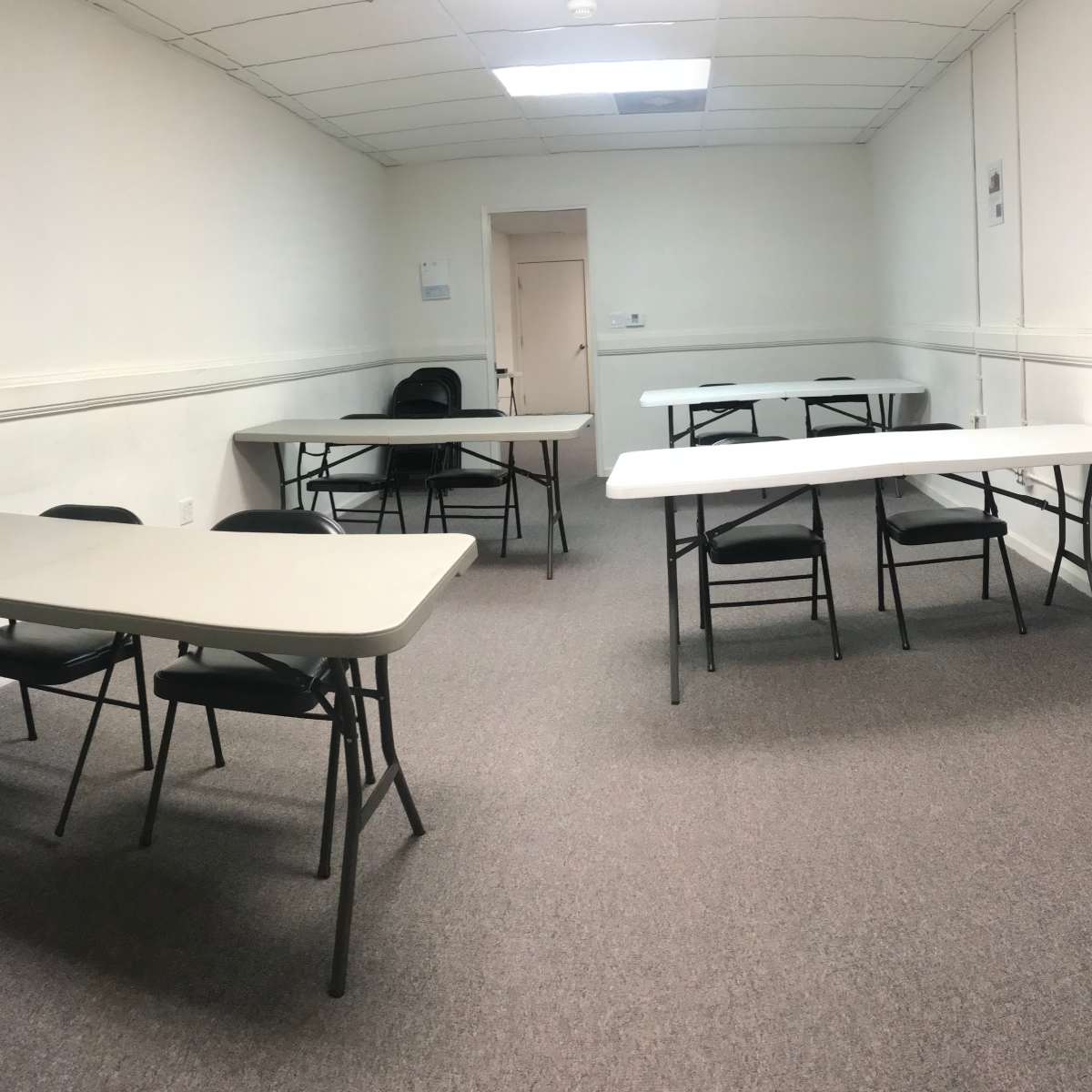 A rectangular room with two sets of folding tables and chairs arranged in a sparse layout against white walls and a beige door.