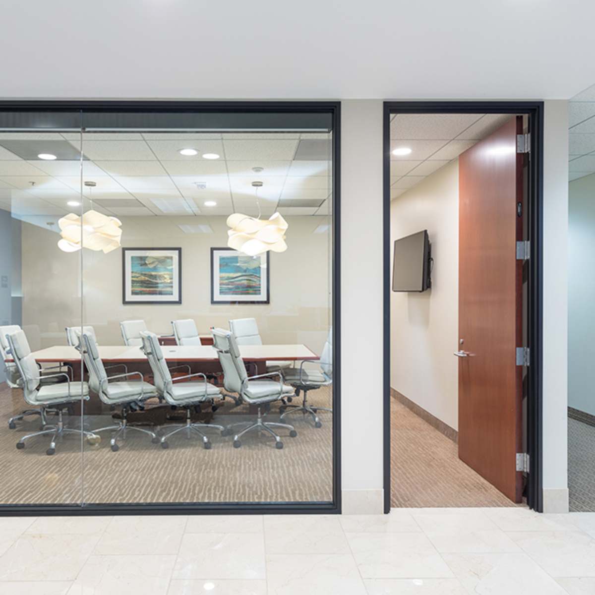 A glass-walled conference room featuring a long table surrounded by chairs is visible through an open door in an office hallway.
