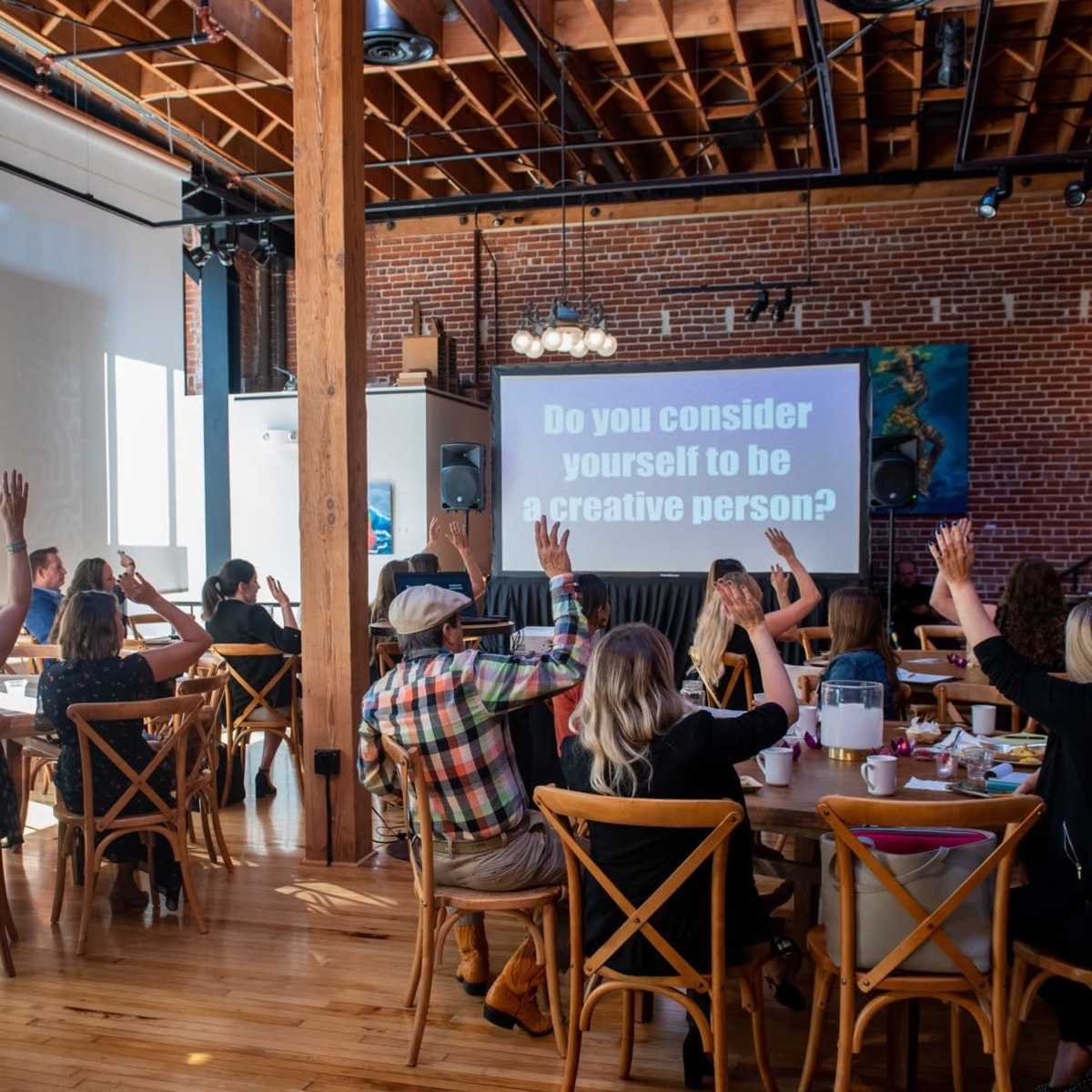 A group of people sits at tables in a spacious room with brick walls, raising their hands in response to a question displayed on a screen.