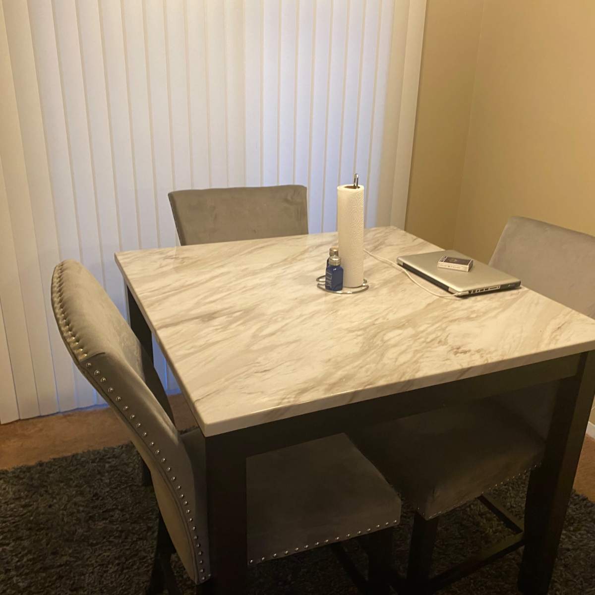 A square dining table with four upholstered chairs is set up in a room with beige walls and vertical blinds.