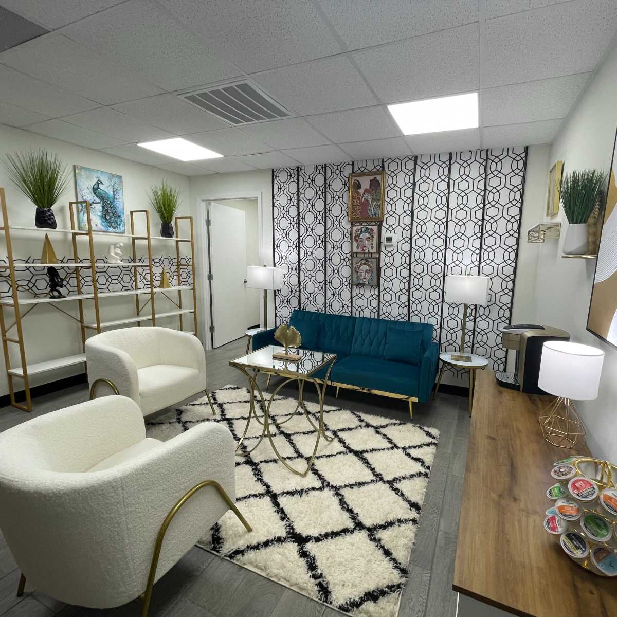 A modern waiting room featuring a teal sofa, two white chairs, a geometric rug, and decorative shelves with plants and artwork.