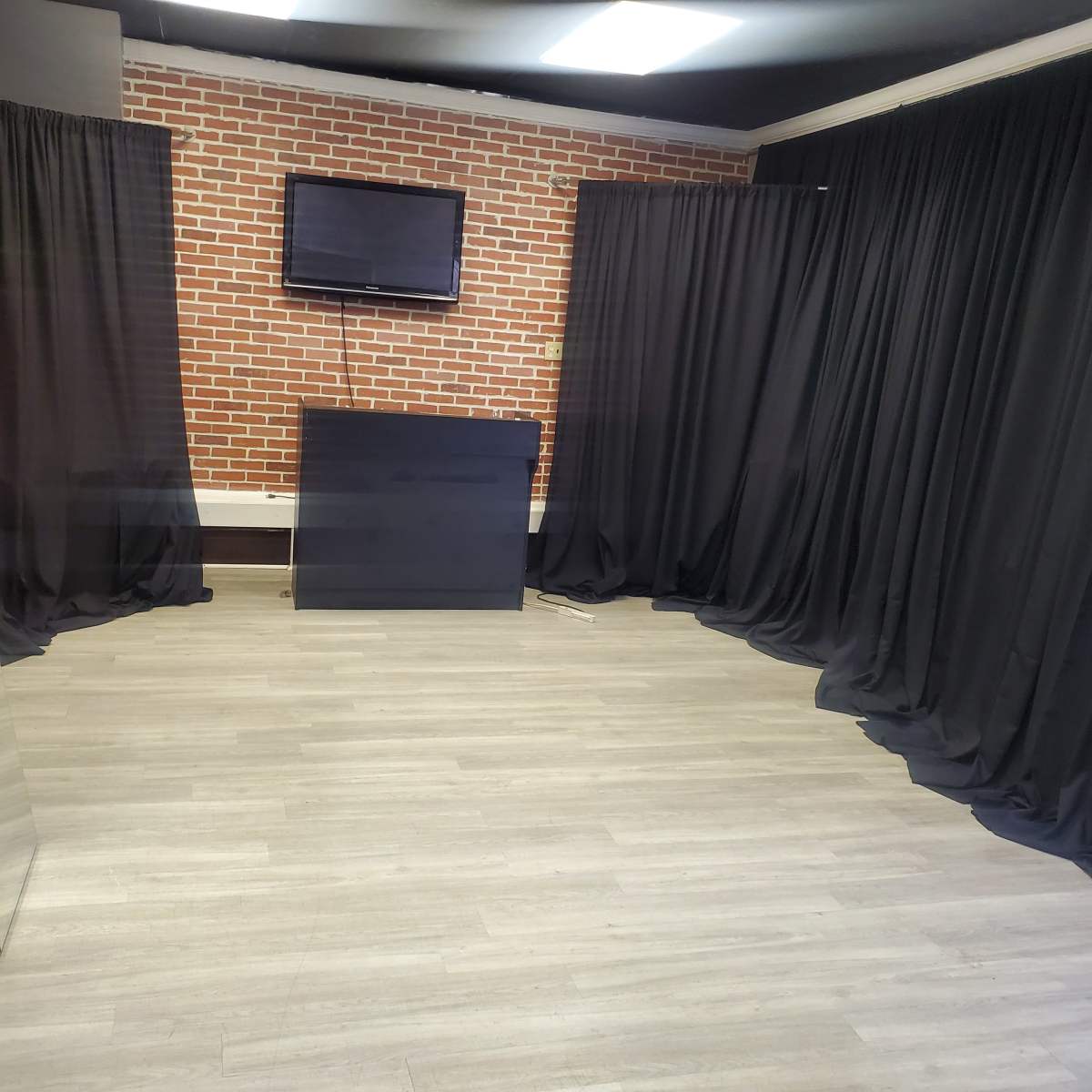A corner of a room with a brick wall, a black screen on the wall, a black counter, and floor covered with light-colored wood flooring, flanked by black curtains.