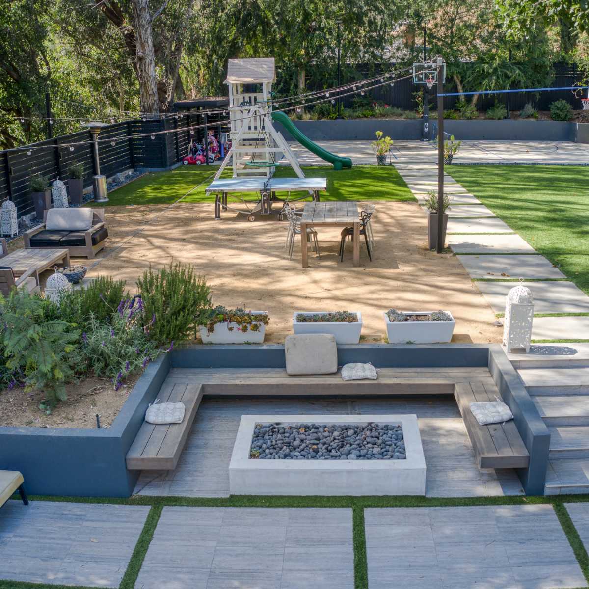 A landscaped outdoor space featuring a lawn, a playground structure, seating areas, and a fire pit.