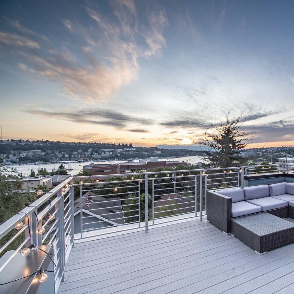 A modern rooftop terrace features a sectional sofa and a coffee table, overlooking a scenic view of the water and landscape at sunset.