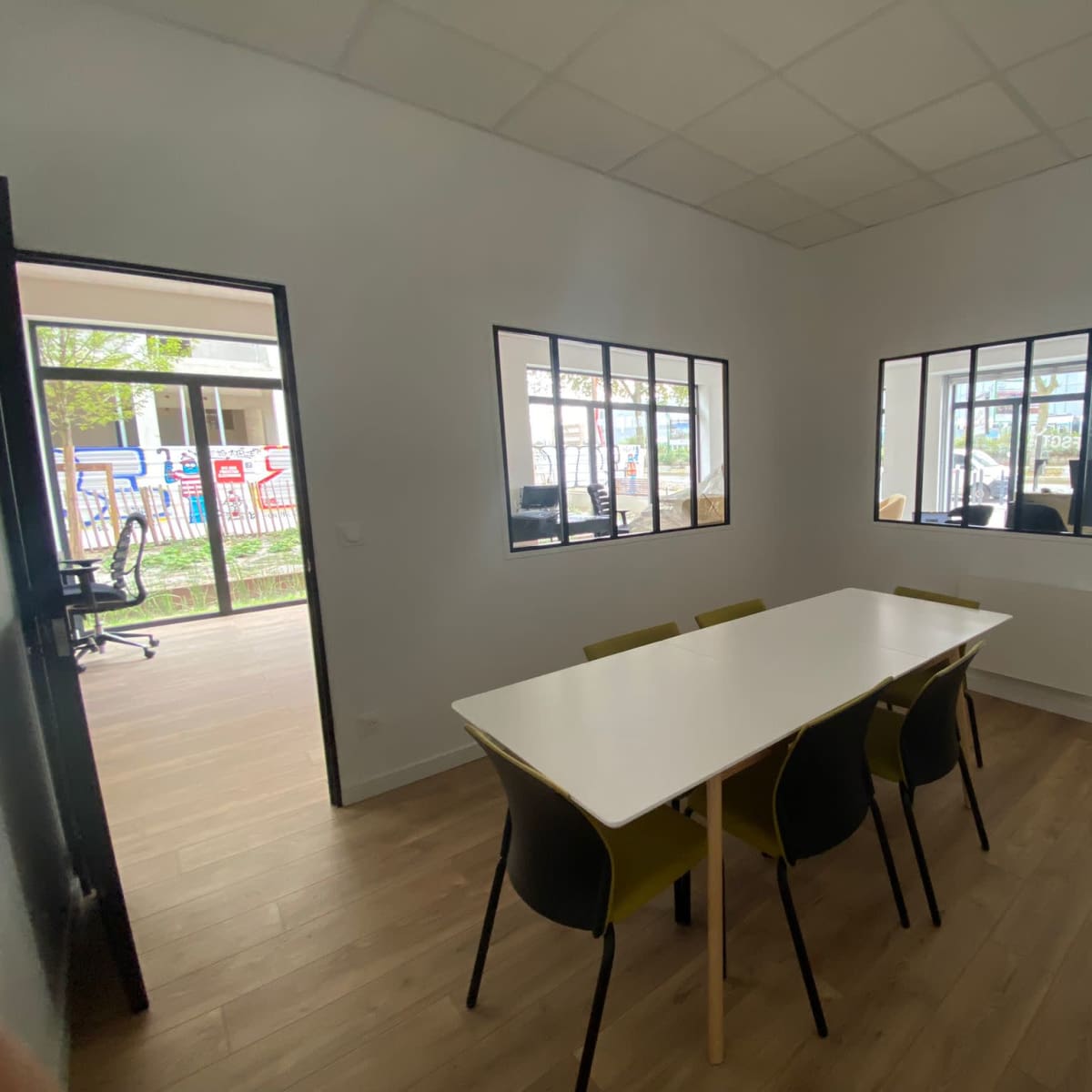 A brightly lit meeting room features a white table surrounded by four green chairs, with large windows overlooking a courtyard.