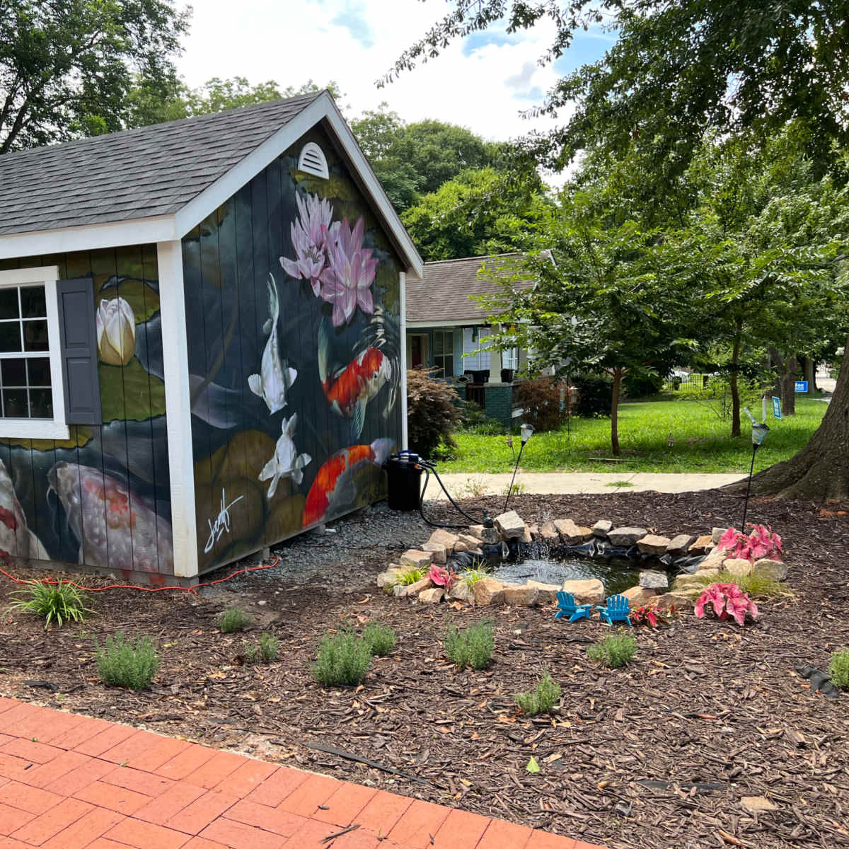 A shed features a mural of koi fish and water lilies, surrounded by a landscaped garden with flowers and stones.