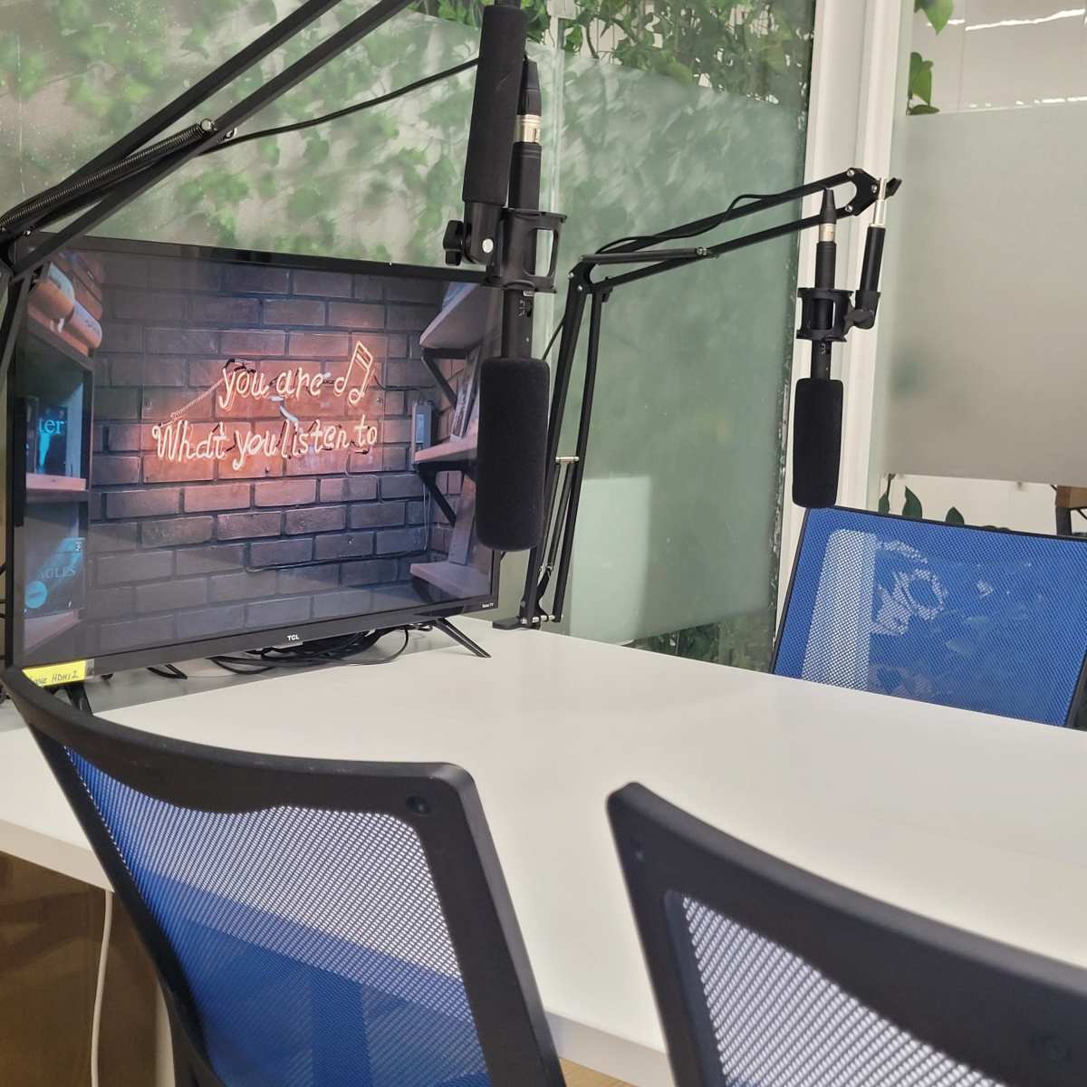 A modern workspace with a table, two blue chairs, and a screen displaying a neon sign that reads "You are what you listen to," along with two mounted microphones.
